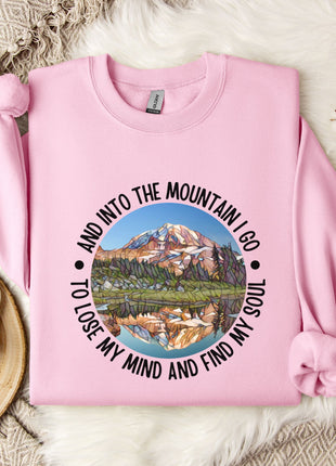 A pink sweatshirt with a mountain landscape design and the text "And into the mountain I go to lose my mind and find my soul" printed on it, along with a mug and some flowers on a bed.