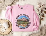 A pink sweatshirt with a mountain landscape design and the text "And into the mountain I go to lose my mind and find my soul" printed on it, along with a mug and some flowers on a bed.