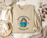 a beige sweatshirt with a graphic design depicting a dumpster with flames coming out of it, accompanied by the text "Emotional Support Dumpster Fire".