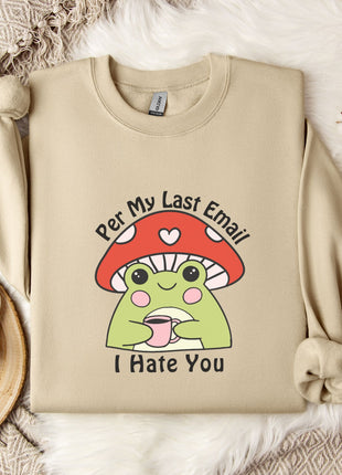 a beige sweatshirt with a cartoon frog wearing a red mushroom hat and the text "Per My Last Email I Hate You" printed on it. The sweatshirt is placed on a bed with a white blanket and a mug, and there are some white flowers nearby.
