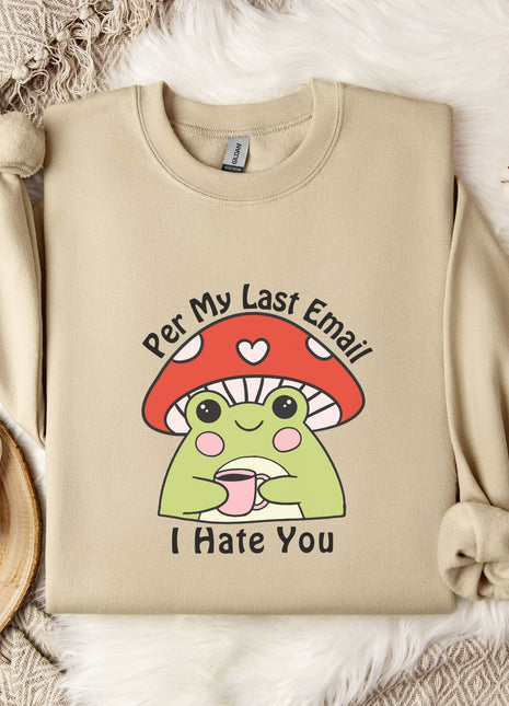 a beige sweatshirt with a cartoon frog wearing a red mushroom hat and the text "Per My Last Email I Hate You" printed on it. The sweatshirt is placed on a bed with a white blanket and a mug, and there are some white flowers nearby.