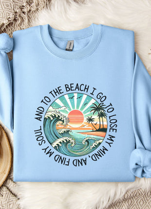 A light blue sweatshirt with a graphic design depicting a beach scene, including a sunset, palm trees, and waves, along with the text "And to the beach I go, I lose my soul, and I find my mind".