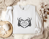A white sweatshirt with a graphic design and text printed on it, along with a mug, a branch of cotton bolls, and a wooden tray with snowflakes on it, all arranged on a white fur rug.