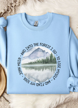 A light blue sweatshirt with a circular design featuring a mountain landscape and a lake, along with the text "AND INTO THE FOREST I GO - TO LOSE MY SON - SO I FIND MY FONDNESS" printed on it. The sweatshirt is laid out on a bed with a white blanket and a mug, and there are some branches and snowflakes in the background.