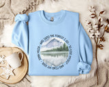 A light blue sweatshirt with a circular design featuring a mountain landscape and a lake, along with the text "AND INTO THE FOREST I GO - TO LOSE MY SON - SO I FIND MY FONDNESS" printed on it. The sweatshirt is laid out on a bed with a white blanket and a mug, and there are some branches and snowflakes in the background.