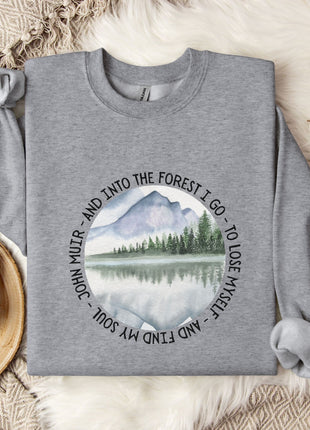 a gray sweatshirt with a circular design on it, depicting a mountain landscape with a lake and pine trees. The design also includes text that reads "And into the forest I go - to lose myself and find my soul". The sweatshirt is laid out on a bed with a white blanket, and there are some cotton balls and a mug nearby.
