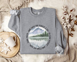 a gray sweatshirt with a circular design on it, depicting a mountain landscape with a lake and pine trees. The design also includes text that reads "And into the forest I go - to lose myself and find my soul". The sweatshirt is laid out on a bed with a white blanket, and there are some cotton balls and a mug nearby.