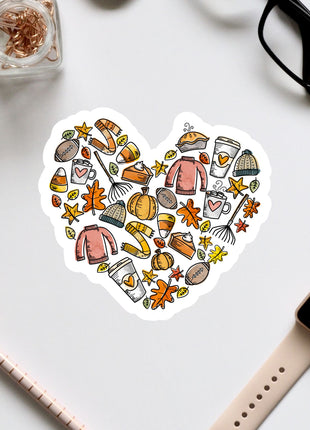 A heart-shaped sticker with various fall-themed items such as pumpkins, leaves, and sweaters, along with a pair of glasses and a watch, all arranged on a white background.