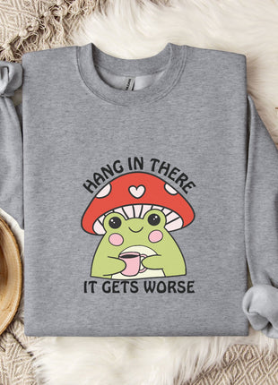 A gray sweatshirt with a mushroom graphic and the text "HANG IN THERE IT GETS WORSE" printed on it, along with a mug and some white flowers on a bed.