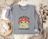 A gray sweatshirt with a mushroom graphic and the text "HANG IN THERE IT GETS WORSE" printed on it, along with a mug and some white flowers on a bed.