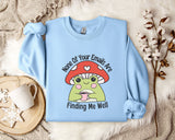 a light blue sweatshirt with a cartoon frog wearing a red mushroom hat and holding a cup, accompanied by the text " none of your emails are finding me well".