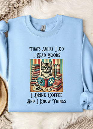 A light blue sweatshirt with a printed design of a cat surrounded by books and a cup of coffee, along with text that reads "Thats what I do I read books I drink coffee and I know things".