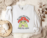 A white sweatshirt with a mushroom character and text printed on it, placed on a bed with a white blanket and a mug.