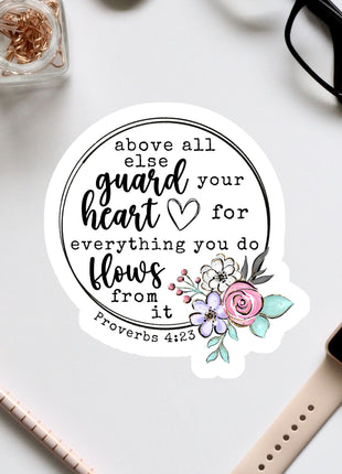 a sticker with a Bible verse and a floral design, surrounded by various office supplies such as a watch, pen, and glasses.