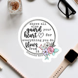 a sticker with a Bible verse and a floral design, surrounded by various office supplies such as a watch, pen, and glasses.