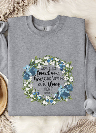 A gray sweatshirt with a floral wreath design and a quote printed on it, along with a mug and some cotton balls on a white fur rug.