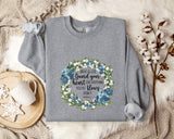 A gray sweatshirt with a floral wreath design and a quote printed on it, along with a mug and some cotton balls on a white fur rug.