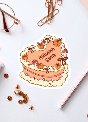 a heart-shaped sticker with the text "Autumn Girl" and various autumn-themed elements, such as pumpkins and bows, surrounding it. The sticker is placed on a white surface, along with a pair of glasses, a pen, a notebook, and some office supplies like a stapler and a pen holder.