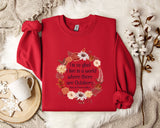 A red sweater with a floral design and a quote printed on it, along with a mug, a white mug, and some white flowers on a white fur rug.
