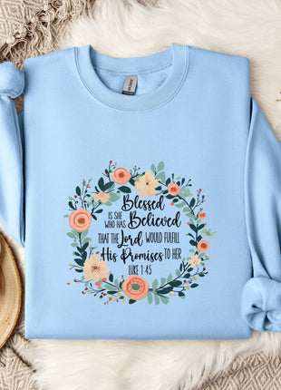 A light blue sweatshirt with a floral wreath design and text printed on it, placed on a bed with a white blanket and a white mug.