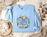 A light blue sweatshirt with a floral wreath design and text printed on it, placed on a bed with a white blanket and a white mug.
