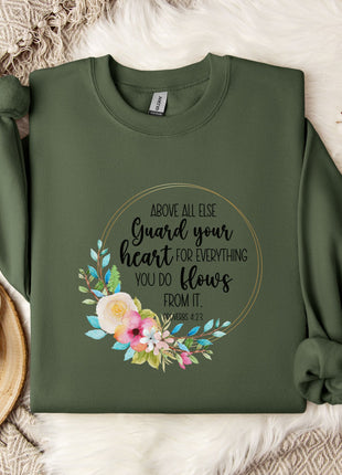 a green sweatshirt with a floral design and text printed on it, along with a mug and a white flower arrangement on a bed.