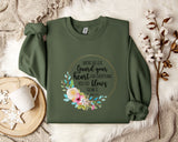 a green sweatshirt with a floral design and text printed on it, along with a mug and a white flower arrangement on a bed.