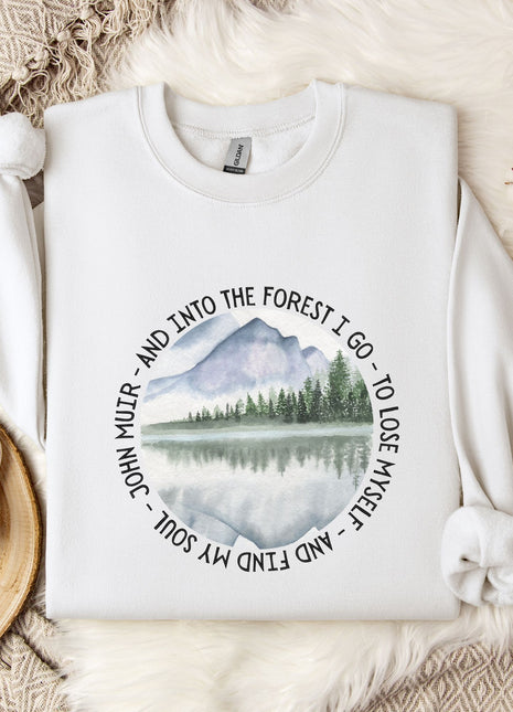 A white sweatshirt with a circular design featuring a mountain landscape and a lake, along with the text "John Muir - And Into the Forest I Go - To Lose My Self - And My Love" printed on it. The sweatshirt is placed on a bed with a white blanket and a mug, and there are some branches and snowflakes in the background.