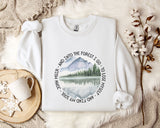 A white sweatshirt with a circular design featuring a mountain landscape and a lake, along with the text "John Muir - And Into the Forest I Go - To Lose My Self - And My Love" printed on it. The sweatshirt is placed on a bed with a white blanket and a mug, and there are some branches and snowflakes in the background.