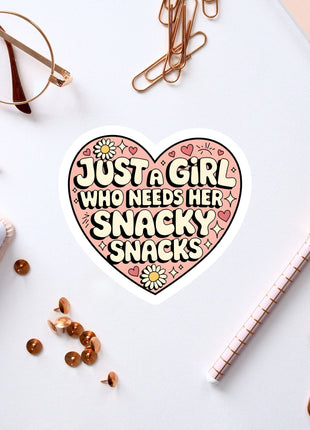 a heart-shaped sticker with the text "Just a girl who needs her snacky snacks" surrounded by various office supplies such as a pen, a pair of glasses, and a notebook.