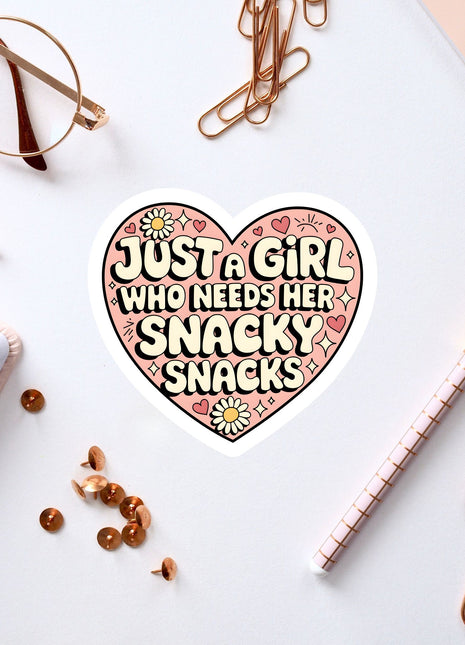 a heart-shaped sticker with the text "Just a girl who needs her snacky snacks" surrounded by various office supplies such as a pen, a pair of glasses, and a notebook.