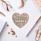 a heart-shaped sticker with the text "Just a girl who needs her snacky snacks" surrounded by various office supplies such as a pen, a pair of glasses, and a notebook.