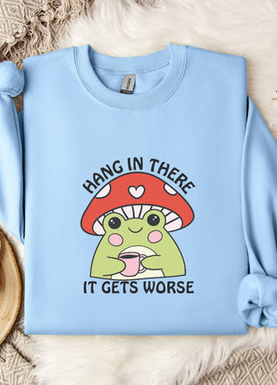 A light blue sweatshirt with a mushroom graphic and the text "HANG IN THERE IT GETS WORSE" printed on it, along with a mug and some flowers on a bed.