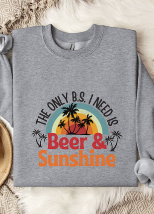 a gray sweatshirt with a graphic design that says "The only B.S. I need is Beer & Sunshine" along with a palm tree and sunset design. The sweatshirt is laid out on a bed with a blanket and a mug, and there are some cotton balls and a branch of flowers nearby.