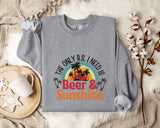 a gray sweatshirt with a graphic design that says "The only B.S. I need is Beer & Sunshine" along with a palm tree and sunset design. The sweatshirt is laid out on a bed with a blanket and a mug, and there are some cotton balls and a branch of flowers nearby.