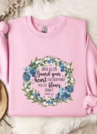 A pink sweatshirt with a floral wreath design and a Bible verse printed on it, along with a mug, a white mug, and some white flowers on a white fur rug.