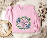 A pink sweatshirt with a floral wreath design and a Bible verse printed on it, along with a mug, a white mug, and some white flowers on a white fur rug.