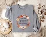 A gray sweatshirt with a floral wreath design and a quote printed on it, placed on a beige blanket with a white mug and a branch of cotton balls nearby.