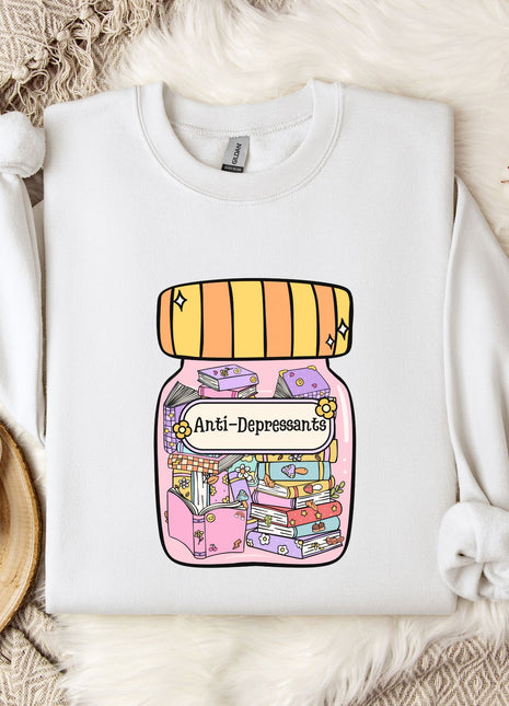 A white sweatshirt with a colorful graphic design depicting a jar with various objects inside, including books and a flower, along with the text "Anti-Depressants" printed on it. The sweatshirt is laid out on a bed with a white blanket and a mug, and there are some flowers nearby.