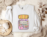 A white sweatshirt with a colorful graphic design depicting a jar with various objects inside, including books and a flower, along with the text "Anti-Depressants" printed on it. The sweatshirt is laid out on a bed with a white blanket and a mug, and there are some flowers nearby.