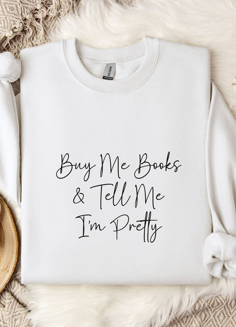 a white sweatshirt with the text "Buy Me Books & Tell Me I'm Pretty" printed on it, along with a mug and some flowers on a bed.