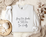 a white sweatshirt with the text "Buy Me Books & Tell Me I'm Pretty" printed on it, along with a mug and some flowers on a bed.