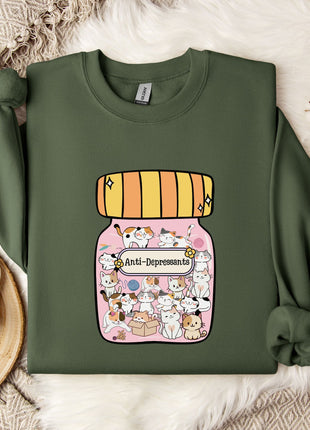 a green sweatshirt with a colorful graphic of a jar filled with various cartoon animals, including cats and dogs, on the front. The sweatshirt is laid out on a bed with a white blanket and a small white mug. There are also some white flowers and a white snowflake visible in the background.