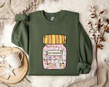 a green sweatshirt with a colorful graphic of a jar filled with various cartoon animals, including cats and dogs, on the front. The sweatshirt is laid out on a bed with a white blanket and a small white mug. There are also some white flowers and a white snowflake visible in the background.
