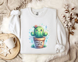 a white sweatshirt with a colorful illustration of a cactus on it, along with a mug and some decorative elements such as a snowflake and a branch of cotton flowers.