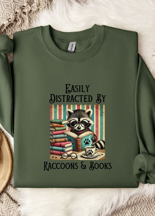 a green sweatshirt with a graphic design of a raccoon reading a book, accompanied by the text "Easily Distracted By Raccoons & Books".