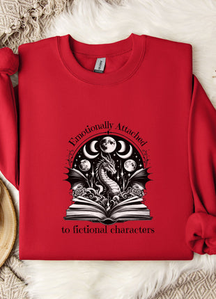 A red sweater with a graphic design of a dragon and a book, along with the text "Emotionally Attached to Fictional Characters" printed on it, is displayed on a bed with a white blanket and a white mug.