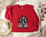 A red sweater with a graphic design of a dragon and a book, along with the text "Emotionally Attached to Fictional Characters" printed on it, is displayed on a bed with a white blanket and a white mug.