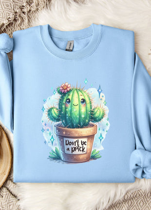 A light blue sweatshirt with a cactus graphic and the text "Don't be a prick" printed on it, along with a mug and some decorative items on a bed.