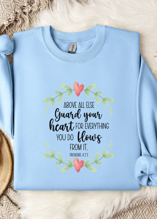 A light blue sweatshirt with a printed Bible verse and a floral design, along with a mug and a branch of cotton flowers, all placed on a white fur rug.